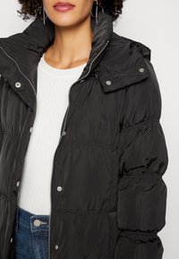 Black puffer jacket with a high collar, zip closure, elasticized sleeves, and button details, worn over a ribbed white sweater.