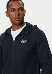 Felpa con zip blu navy, dotata di tasca frontale, cappuccio regolabile con cordini e logo EA7. Realizzata in tessuto morbido con una texture liscia.
