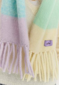 Miękki wełniany szalik z pastelowymi blokami kolorów w odcieniach fioletu, niebieskiego, żółtego i zielonego, z frędzlami na końcach i małą fioletową metką z tkaniny.