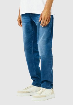 Jeans de mezclilla azul con un corte recto, lavado medio, desvanecimientos sutiles, detalles en los bolsillos, combinados con zapatillas blancas y beige.