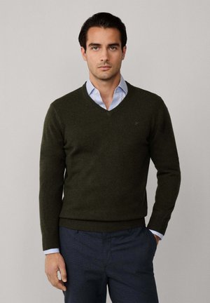 Pull en laine vert olive avec un col en V, des poignets et une taille côtelés, porté sur une chemise col bleu clair. Logo simple sur la poitrine.