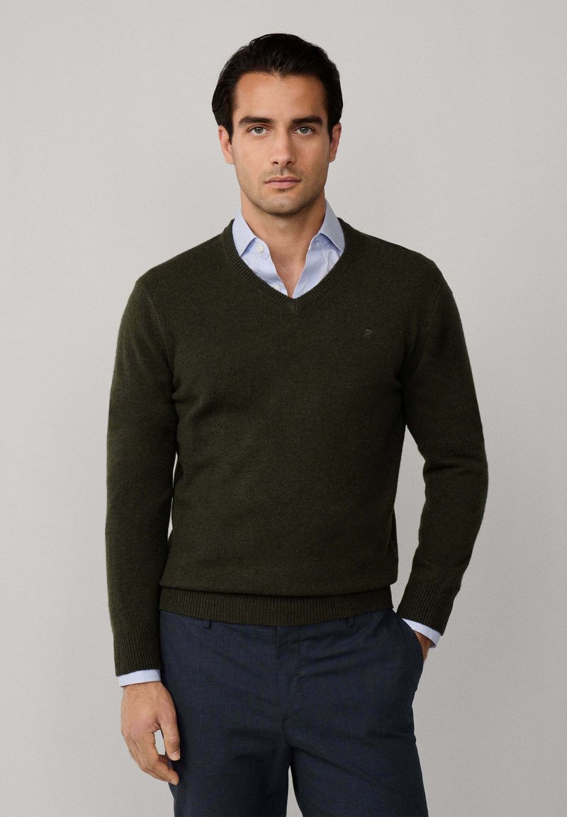 Olivgrön ulltröja med V-ringad design, ribbade ärmslut och midja, över en ljusblå skjorta med krage. Enkelt logotyp på bröstet.