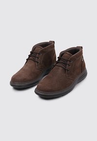 Botas de tobillo de ante marrón con cordones negros, cuellos acolchados y suelas de goma negras, situadas sobre un fondo gris claro.