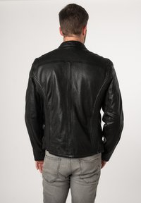 Giacca in pelle nera con un design aderente, superficie texture e dettagli delle cuciture decorativi. Indossata sopra pantaloni grigi, vista da dietro.