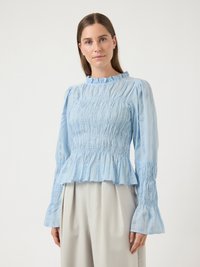 Νεαρή γυναίκα που φοράει ανοιχτό γαλάζιο μπλούζα με ρυτίδες και μακριά μανίκια, καθώς και μπεζ πλισέ παντελόνια, στέκεται μπροστά σε λευκό φόντο.