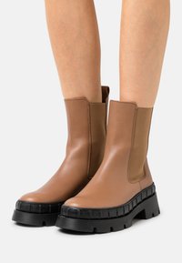 Brązowe skórzane botki za kostkę z masywną czarną gumową podeszwą, elastycznymi panelami po bokach i gładką fakturą. Charakteryzują się zaokrąglonym noskiem i solidną konstrukcją.