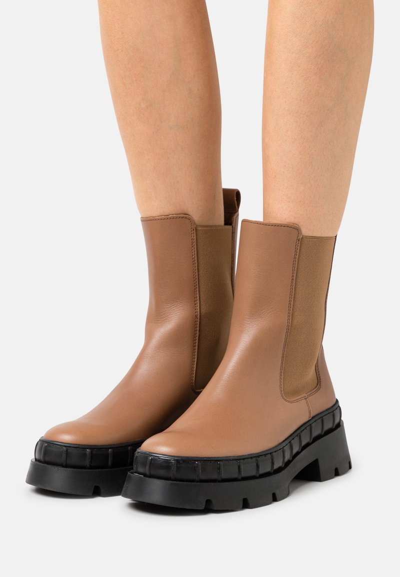 Brązowe skórzane botki za kostkę z masywną czarną gumową podeszwą, elastycznymi panelami po bokach i gładką fakturą. Charakteryzują się zaokrąglonym noskiem i solidną konstrukcją.