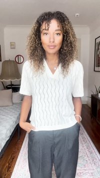 Top in maglia bianco, a maniche corte con scollatura a V e motivo ondulato; abbinato a pantaloni scuri, larghi. Sullo sfondo ci sono arredamenti morbidi.