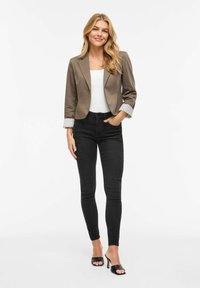 Jeune femme portant un blazer marron court, un haut blanc, un jean skinny noir et des sandales à talons noires, debout avec une main dans la poche.