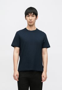 Tmavě modré tričko s krátkým rukávem z bavlněné látky, s kulatým výstřihem a volnějším střihem. Nošeno s černými kalhotami.