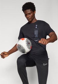 Crna Nike majica s plavim tekstom i logotipom, crne atletske hlače s bijelim logotipom, igrač izvodi trik s šarenom nogometnom loptom.
