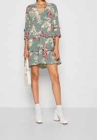 Robe florale verte à manches longues, avec des volants et un décolleté en V. Le motif est orné de fleurs roses et de papillons. Associée à des talons bloc blancs.