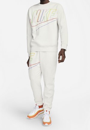 Personne portant un sweat-shirt Nike blanc et un pantalon de jogging assorti avec un logo multicolore en contour, assortis à des baskets Nike blanches et orange.