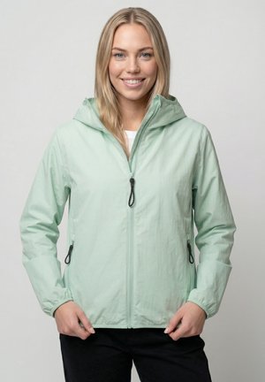 Lächelnde Frau in einer hellgrünen, zip-up Kapuzenjacke und schwarzen Hosen, stehend vor einem schlichten grauen Hintergrund.