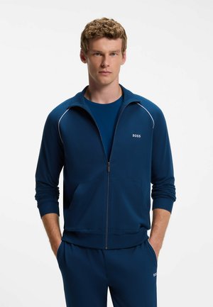 Junger Mann mit lockigem blondem Haar trägt eine marineblaue BOSS Reißverschlussjacke und passende Hose, die Hände in den Taschen, vor weißem Hintergrund.