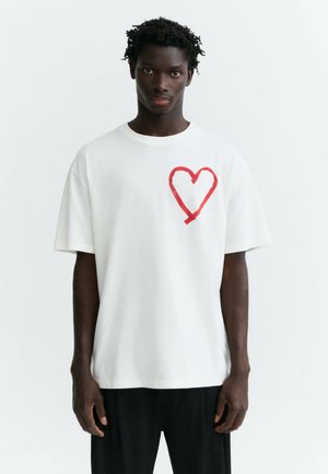 Giovane uomo con capelli corti e ricci che indossa una t-shirt bianca con un grande cuore rosso sul petto sinistro e pantaloni neri.