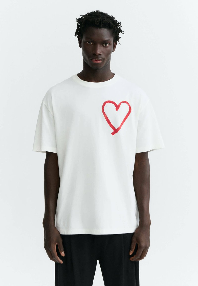 Jeune homme aux cheveux courts et bouclés portant un t-shirt blanc avec un grand cœur rouge sur la poitrine gauche et un pantalon noir.