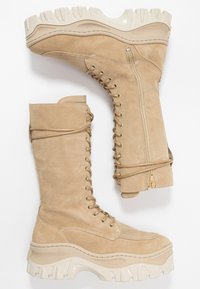 Botas de ante beiges hasta el tobillo y hasta la rodilla con suela de goma blanca gruesa, cordones en la parte delantera, cremallera lateral y detalles decorativos de cordones.