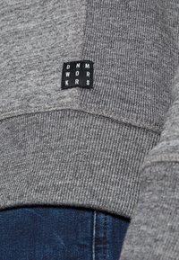 Grijze sweatshirt met ribbeltextuur, voorzien van een zwart label met witte tekst "DNM WORKRS." Close-up toont de stikseldetails en de consistentie van de stof.