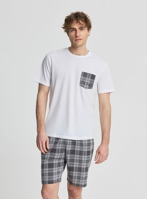 Młody mężczyzna w białej koszuli z krótkim rękawem, z szarą kratką na kieszeni i pasujących krótkich szortach w szarą kratę, stoi na jednolitym tle.