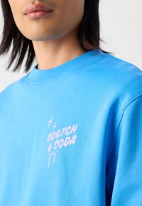 Blauwe crewneck sweatshirt gemaakt van een zachte stof. Het heeft een roze logo "SCOTCH & SODA" met druppelende accenten aan de linkerkant.