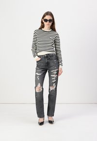 Black and white striped sweater, high-waisted ripped black jeans, and black patent shoes. Model wearing sunglasses, standing against a plain background.