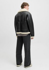 Ung mand står oprejst, iført sort shearling-jakke, ternet skjorte, løse sorte jeans og hvide sneakers, med ryggen til en ensfarvet baggrund.