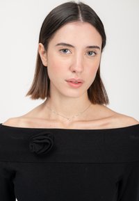 Schwarzes Off-Shoulder-Oberteil aus strukturiertem Stoff, mit einer rosenförmigen Akzentuierung am Ausschnitt. Schlichte silberne Halskette mit einem kleinen Anhänger.