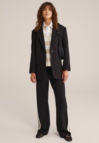 Blazer noir oversize, chemise blanche à col, pull rayé beige, pantalon noir ample avec accents blancs, et chaussures en mailles noires.