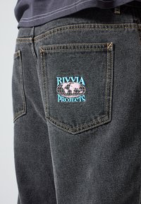 Μαύρα τζιν από τζιν ύφασμα με κεντητό σχέδιο σε τσέπη στην πλάτη σε ανοιχτό μπλε και ροζ, με τη φράση "RIVIA PROJECTS" πάνω από ένα σχέδιο γης.