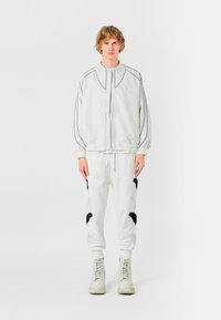 Chaqueta blanca con cremallera y contornos negros contrastantes, combinada con pantalones blancos que presentan acentos bordados en negro. El modelo lleva botas blancas.