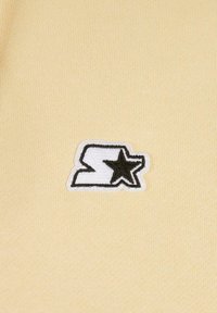 Un tessuto giallo con una consistenza morbida presenta un logo ricamato in bianco e nero, che mostra una "S" stilizzata con un accento a forma di stella.