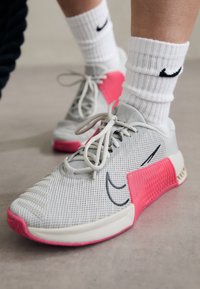 Zapatillas deportivas de color gris claro con parte superior de malla, acentos rosas en el talón y el swoosh negro de Nike. Los cordones son grises y texturizados.