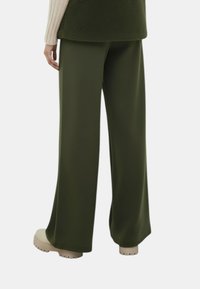 Wijde broekspijpen in donkergroen stof, met een gladde textuur en een ontspannen pasvorm, gecombineerd met lichte schoenen.