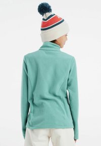 Pullover in pile blu chiaro con colletto alto e maniche lunghe. Indossato con un cappello bianco con pompon e dettagli a strisce colorate.
