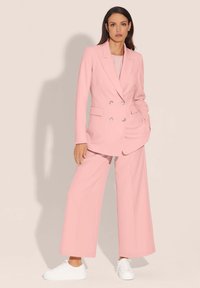 Roze pantalonpak met een dubbelzijdige blazer en brede broekspijpen. Glad stof, twee frontzakken, gecombineerd met witte sneakers.