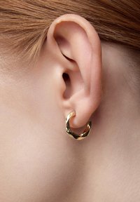 Złota kolczyk w kształcie obręczy z przekręconym wzorem oraz teksturowaną powierzchnią. Znajduje się w płatku ucha obok gładkiej skóry.