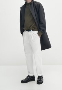 Cappotto nero, maglione a coste verde oliva, pantaloni bianchi, scarpe nere. Design piatto, texture liscia, vestibilità strutturata e dettagli con bottoni sul cappotto.