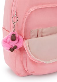 Růžový batoh s hladkým textilním povrchem; obsahuje kapsu se zipem. Připojené je chlupaté růžové klíčenka gorily s hnědými akcenty.