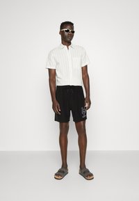 White and striped short-sleeve shirt, black shorts with graphic detail, gray sandals, and sunglasses. Casual summer outfit.