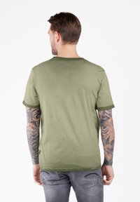 Hombre con brazos tatuados vistiendo una camiseta verde oliva y jeans grises, de pie de espaldas contra un fondo blanco liso.