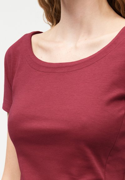 Γυναίκα που φοράει εφαρμοστό βυσσινί τοπ με κοντά μανίκια και στρογγυλή, φαρδιά λαιμόκοψη, με φόντο απλή επιφάνεια.