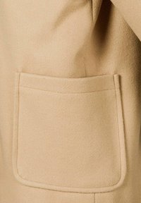 Béžový kabát z vlnenej zmesi s jemnou textúrou, ktorý má na prednej strane obdĺžnikovú náprsníkovú kapsu pre funkčný detail.