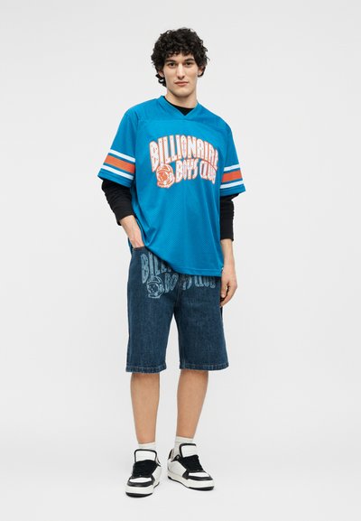 Νεαρός άνδρας φοράει μπλε αθλητική φανέλα «Billionaire Boys Club», τζιν σορτς με λογότυπο, μακρυμάνικη μαύρη μπλούζα, λευκές κάλτσες και ασπρόμαυρα αθλητικά παπούτσια.