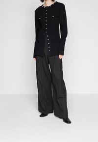 Mujer vistiendo un cárdigan negro con botones y dos bolsillos frontales, combinado con pantalones negros de pierna ancha y zapatos negros, sobre un fondo liso.