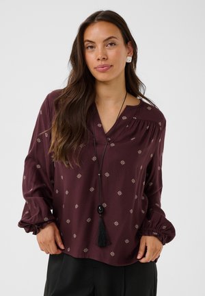 Femme aux longs cheveux foncés porte un chemisier ample bordeaux avec de petits motifs, un pantalon noir, un collier à pompons et des boucles d’oreilles carrées en argent.