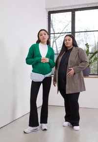 Duas pessoas estão de pé em frente a uma janela. A primeira usa um cardigan verde de malha com padrões, um suéter branco de gola alta, calças pretas largas e ténis. A segunda está vestida com um casaco castanho oversized em padrão xadrez e uma camisola preta com calças. Uma bolsa azul clara está presa à cintura da primeira.