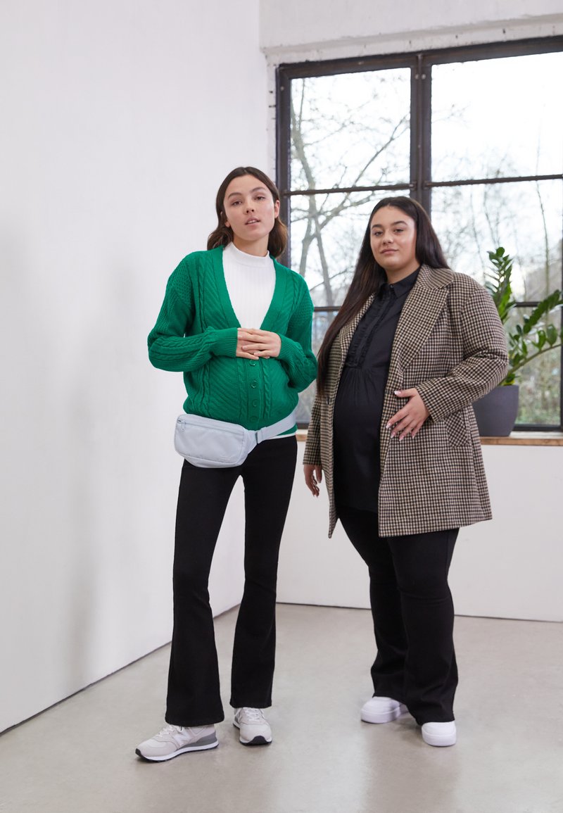 Duas pessoas estão de pé em frente a uma janela. A primeira usa um cardigan verde de malha com padrões, um suéter branco de gola alta, calças pretas largas e ténis. A segunda está vestida com um casaco castanho oversized em padrão xadrez e uma camisola preta com calças. Uma bolsa azul clara está presa à cintura da primeira.