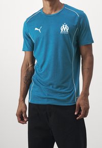 Maglietta sportiva turchese in tessuto morbido con piping bianchi, logo Puma e stemma di Marsiglia con il testo "DROIT AU BUT."