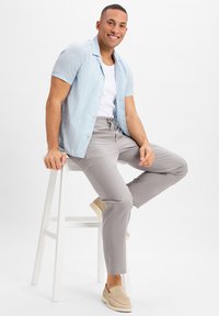 Hellblaues kurzärmliges Hemd mit Palmenmuster, weißes Tanktop, graue Jogginghose mit Zugband und beige Slippers. Modell sitzt auf einem weißen Hocker.